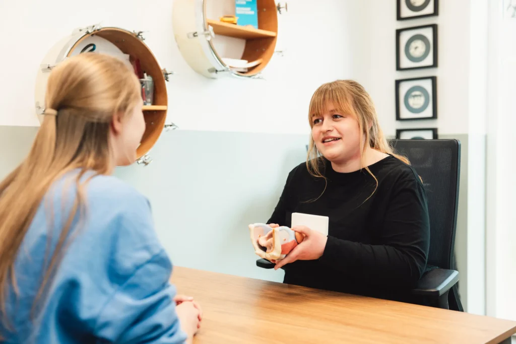 Eine Frau in einem schwarzen Oberteil sitzt an einem Schreibtisch und hält ein medizinisches Kehlkopfmodell in der Hand, während sie einer anderen Frau mit langen blonden Haaren, die ihr in einem Sprechzimmer gegenüber sitzt, etwas erklärt.