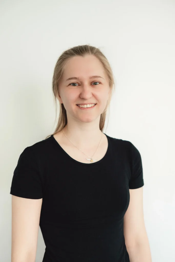 Eine junge Frau mit heller Haut und glattem blondem Haar, die ein schwarzes Kurzarm-Shirt und eine silberne Halskette trägt, steht lächelnd vor einem schlichten weißen Hintergrund.