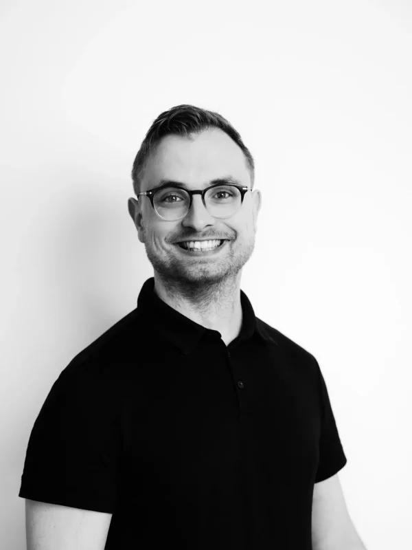 Ein Mann mit Brille und in einem schwarzen Polohemd lächelt vor einem schlichten weißen Hintergrund in die Kamera. Das Foto ist schwarz-weiß.