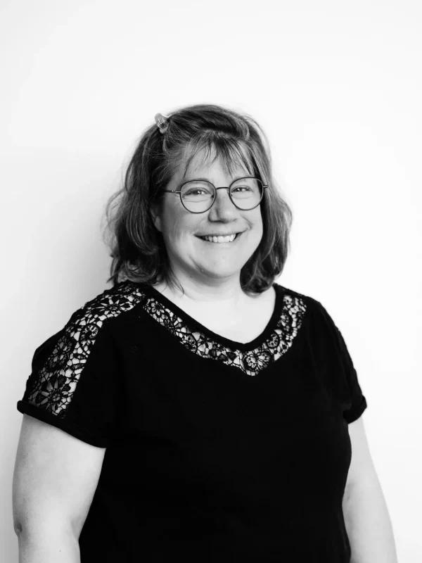 Eine Frau mit schulterlangem Haar, Brille und einer Haarspange lächelt in die Kamera. Sie trägt ein schwarzes Oberteil mit Spitzendetails an den Schultern. Der Hintergrund ist schlicht und weiß. Das Bild ist in Schwarz-Weiß gehalten.