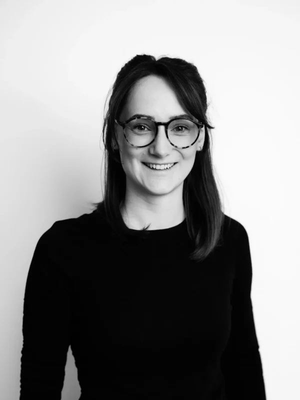 Eine Frau mit glattem, schulterlangem Haar und Brille lächelt in die Kamera. Sie trägt ein langärmeliges, dunkles Oberteil und steht vor einem schlichten, hellen Hintergrund. Das Foto ist in Schwarz-Weiß.