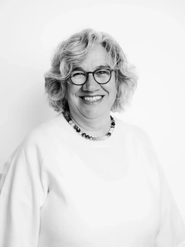 Eine Frau mit kurzen, lockigen Haaren und Brille lächelt in die Kamera. Sie trägt ein helles Oberteil und eine Perlenkette und steht vor einem schlichten weißen Hintergrund. Das Foto ist in Schwarz-Weiß.