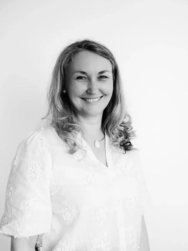 Eine Frau mit gewelltem Haar, die eine helle, bestickte Bluse und eine Halskette trägt, lächelt vor einem schlichten weißen Hintergrund in die Kamera. Das Foto ist in Schwarz-Weiß.