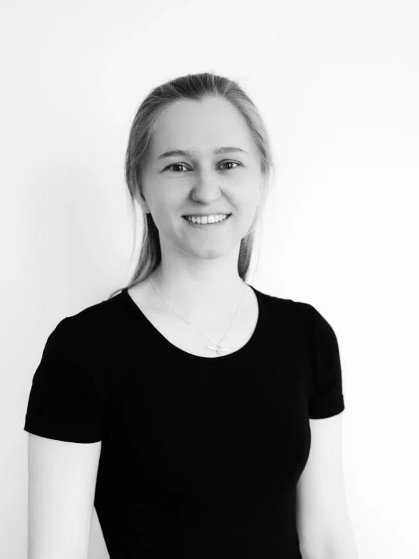Eine junge Frau mit langen, hellen Haaren, die ein schwarzes, kurzärmeliges Shirt und eine Halskette trägt, steht lächelnd vor einem schlichten weißen Hintergrund. Das Foto ist in Schwarz-Weiß.