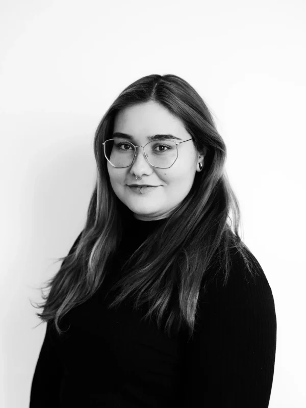 Eine Frau mit langen, glatten Haaren und Brille, die einen schwarzen Rollkragenpullover trägt, steht vor einem schlichten weißen Hintergrund und lächelt sanft in die Kamera. Das Foto ist in Schwarz-Weiß.