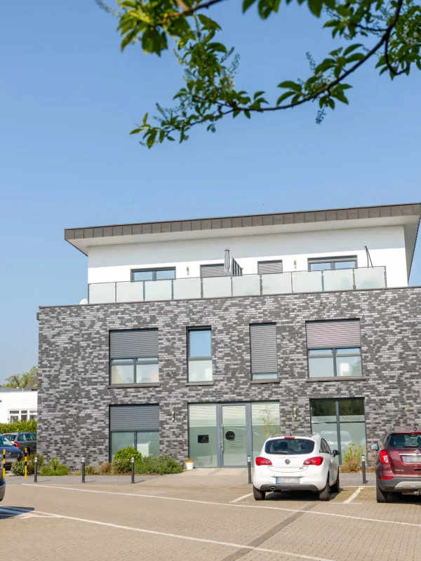 Ein modernes zweistöckiges Gebäude mit einer Fassade aus Ziegeln und weißem Putz, großen Fenstern und einer Dachterrasse. Auf dem Parkplatz davor stehen mehrere Autos, und ein Ast rahmt den oberen Bildrand vor dem Hintergrund eines strahlend blauen Himmels ein.