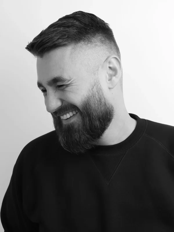 Ein Mann mit kurzem Haarschnitt und Vollbart lächelt und blickt zur Seite; er trägt ein dunkles Sweatshirt vor einem schlichten weißen Hintergrund.