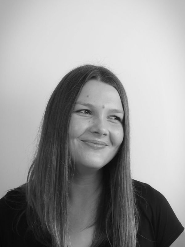 Schwarz-Weiß-Foto einer jungen Frau mit glattem, langem Haar, die sanft lächelt und leicht nach links blickt. Sie trägt ein dunkles Shirt und posiert vor einem schlichten, hellen Hintergrund.